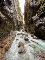 A powerful stream flows through the rocky riverbed of Partnachklamm Gorge, winding between towering cliffs and creating a dramatic, rugged valley scene in the Bavarian Alps.