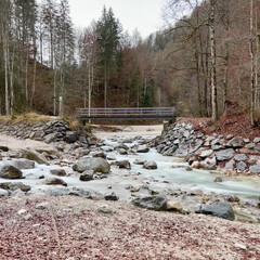 A small bridge near Partnacher Felsentürme spans a clear river, framed by a misty forest and towering mountains, creating a serene, mystical scene at Partnachklamm Gorge.