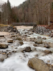 A small bridge near Partnacher Felsentürme spans a clear river, framed by a misty forest and towering mountains, creating a serene, mystical scene at Partnachklamm Gorge.