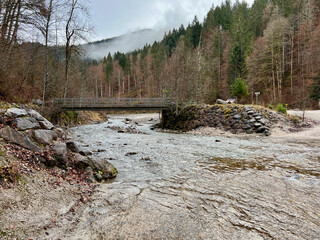 A small bridge near Partnacher Felsentürme spans a clear river, framed by a misty forest and towering mountains, creating a serene, mystical scene at Partnachklamm Gorge.