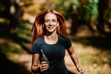 Beautiful young redhead woman walking and jogging in the woods with bottle of drinking water....