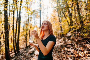 Beautiful young redhead woman walking and jogging in the woods with bottle of drinking water. Healthy lifestyle and sport in nature concept.