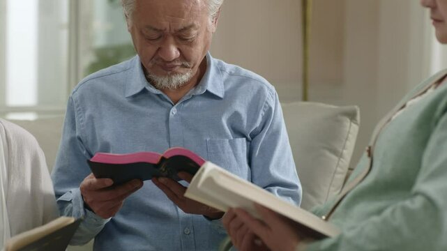 group of senior asian people sitting on family couch reading bible together