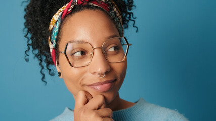 Happy young woman with glasses dressed in blue sweater turns her head posing for camera on blue background in studio