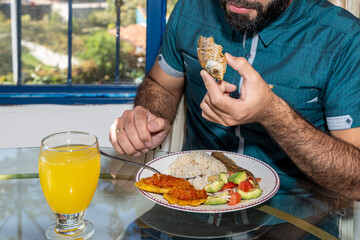 Man with beard at home eating typical Colombian dish, fish with coconut rice, banana and salad with...