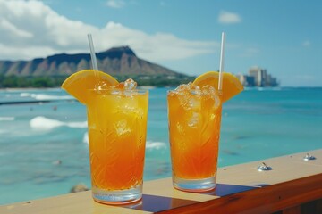 Two glasses of orange juice with straws in them are on a wooden railing overlooking the ocean. The scene is relaxed and peaceful, with the ocean waves in the background