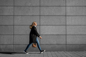 Middle-aged blonde woman in black coat, jeans and sunglasses walking against background of concrete...