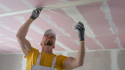 The worker make a plasterboard ceiling. He does taping plaster drywall ceiling joints. He is ...