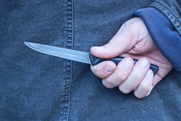 hand of a male criminal in black clothes and a gray knife behind his back on the street