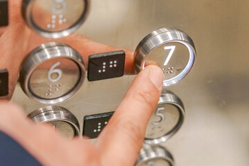 An index finger presses the seventh floor button in an elevator, illuminated with white light, with Braille numbers for visually impaired people, on a gray metallic panel with mirror reflections