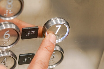 An index finger presses the seventh floor button in an elevator, illuminated with white light, with Braille numbers for visually impaired people, on a gray metallic panel with mirror reflections
