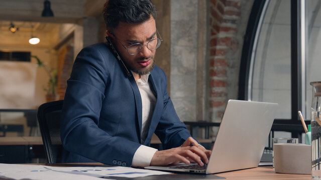 Multitasking businessman stressed busy Indian Arabian business man displeased male office entrepreneur mobile phone call annoyed anxious guy write work laptop pc problem stress deadline at workplace