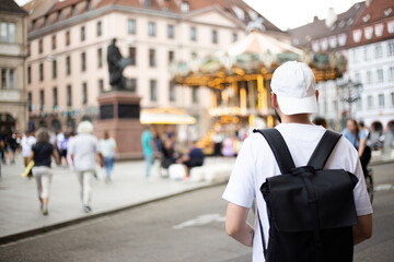 A young man with white cap and black backpack on street of  Carrousel de la Place Gutenberg, Strasbourg. blurred background with walking people. Travel concept and urban lifestyle.