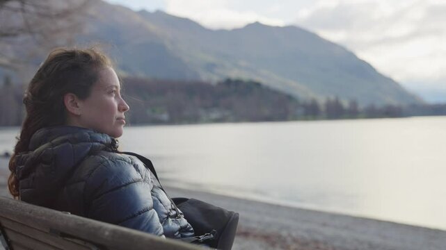 Female tourist or student sitting on bench overlooking beautiful Wanaka Lake. Traveling in New Zealand South Island young woman stopping for relaxation break or rest enjoying amazing picturesque view