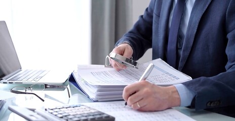 Businessman or auditor is writing notes while using a calculator and magnifying glass while financial documents analyzing. Audit and taxes concept