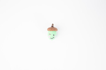 a single acorn with a painted face on white background. one winking eye, a smiling mouth, and pink...