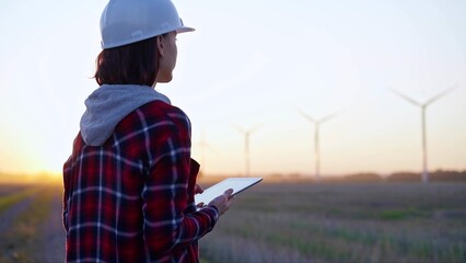 Woman engineer taking notes with a tablet computer on a field with wind turbines, as the sun sets. Concept of clean energy and engineering