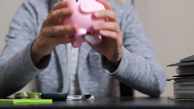 Close-up of man empties a pink piggy bank onto his desk, watching as coins scatter across the surface and counting it, slow motion. Savings, budgeting and financial planning concept