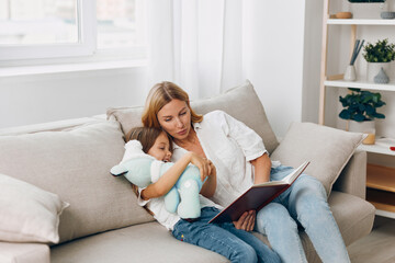 Fototapeta premium Woman reading a book to a little girl on a cozy couch in a warmly lit living room