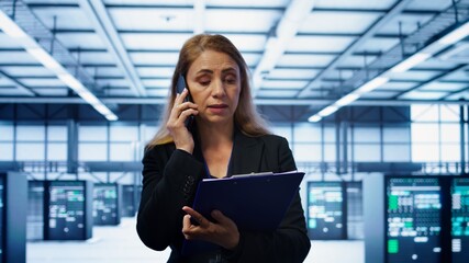 IT specialist looking over data center maintenance checklist, talking on phone in server farm....