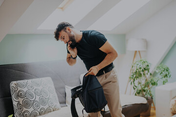 A student in a modern living room packs his bag while talking on the phone, exemplifying...