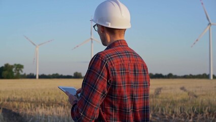 Man engineer at work is wearing a white protective helmet and taking notes with a tablet computer in a field with wind turbines, as the sun sets. Clean energy concept