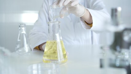 A scientist, wearing a lab coat and white protective gloves, is pouring a yellow oily liquid from one beaker to another in laboratory, close up. Medicine and science concept