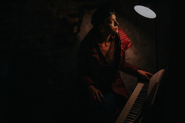 A thoughtfully dressed woman plays piano in a dimly lit room, illuminated by a single lamp. The...