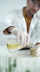 Man scientist with lab coat, white gloves, and protective glasses, is pouring a yellow oily liquid from one beaker to another near microscope in a laboratory, vertical close-up view