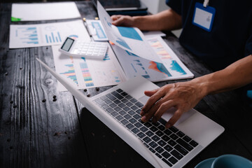 businessman sits at his office desk, analyzing financial documents and graphs on his laptop. paperwork, calculator, and charts, he strategizes investments, audits, tax planning for success.