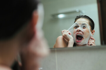 Surprised Asian woman holding a facial mask while looking at her reflection in the bathroom mirror. Skincare and beauty routine concept in a relaxed setting.