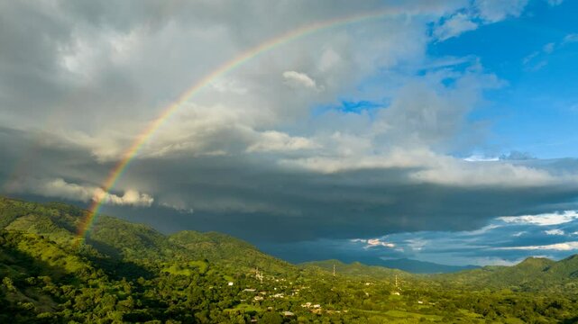 Hyperlapse de un arco&iacute;ris que sale de las monta&ntilde;as sobre un pueblo y se refleja en las nubes oscuras a punto de caer una tormenta