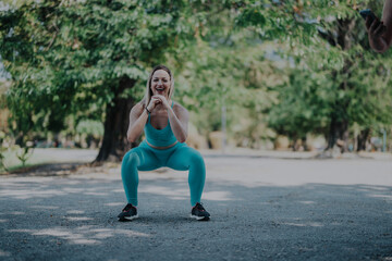 A woman in teal athletic outfit performs a squat in a scenic park, surrounded by greenery,...