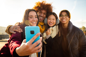 Fototapeta premium Young caucasian female taking selfie with group of multiracial friends outdoors in winter sunny day using mobile phone.