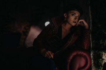 A fashionable woman in a red leather jacket and hat posing on a leather chair, exuding confidence...