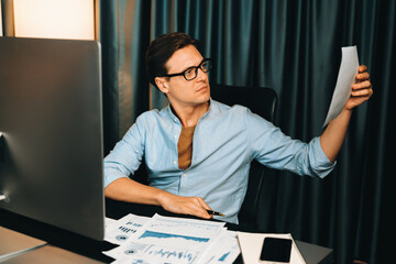 Smart businessman holding paperwork report paragraph dynamic database, looking at pc screen with analyzing global target evaluation value profit company at modern home office at night time. Pecuniary.