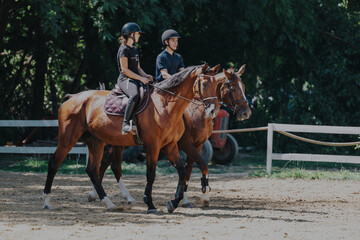 Two young equestrians riding horses on a sunny day in a lush outdoor setting. They are wearing helmets and casual attire, enjoying an active and leisurely horseback session.