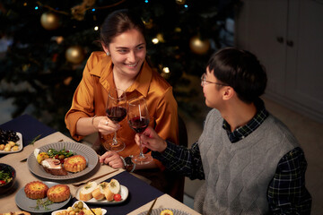 High angle shot of cheerful woman in golden blouse clinking glasses raising toast during Christmas celebrating with male friend, while sitting at dining table set with festive dishes