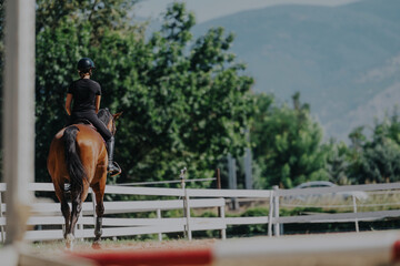 A young equestrian rider enjoys a sunny day riding a horse in an outdoor arena. The setting is...