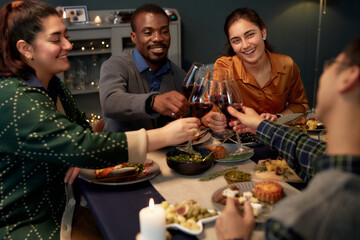 Medium shot of cheerful woman and Black man clinking wine glasses while sitting at festive dining table celebrating Christmas with friends at home party
