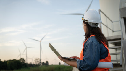 A female engineer  wearing a hard hat and safety vest is looking at a laptop computer. She is...
