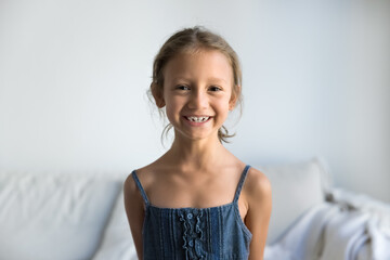 Happy adorable early school aged little girl posing for home portrait in living room, looking at...