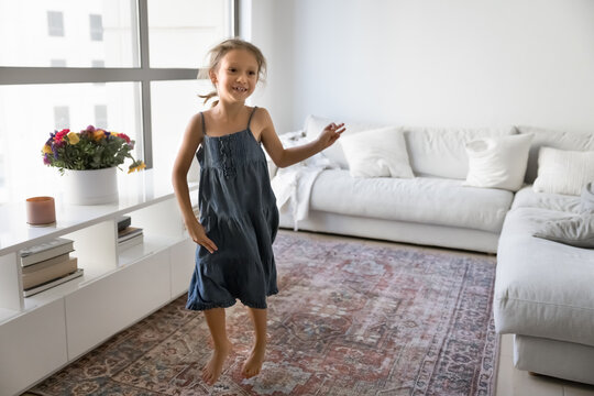 Happy hyperactive little kid girl dancing and jumping to music in home living room, flying over carpeted floor, smiling, laughing, having fun alone, enjoying domestic activity