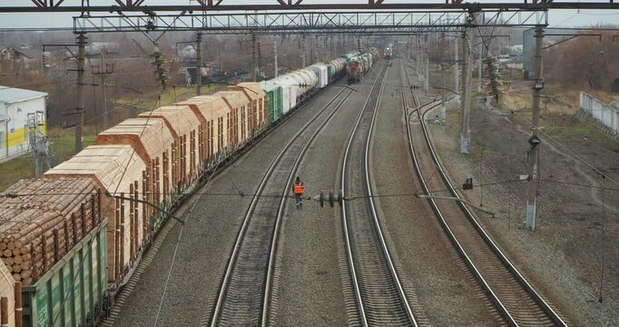 Transport. A road worker in an orange vest walks at the railway station along the railway past a freight train that is loaded with logs and planks. Hand held. Siberia.