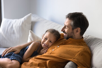 Cheerful loving Hispanic daddy and happy sweet daughter kid resting on comfortable white home couch, looking away, smiling, hugging, enjoying family leisure, laziness, watching TV
