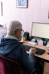 A senior man wearing glasses types on a keyboard while working at a desk with a computer indoors