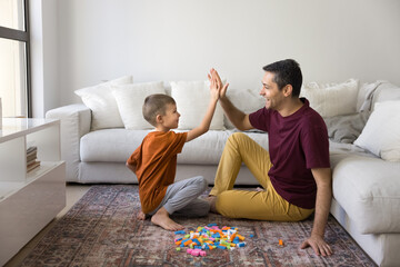 Cheerful father and cute son child clapping hands, giving high five, playing with toy building cubes on carpeted floor at cozy home, sitting at heap of colorful construction blocks, having fun