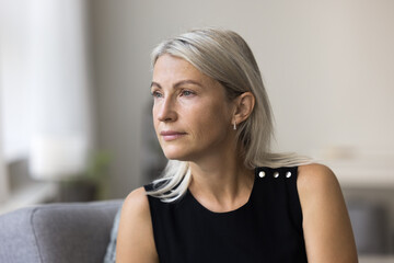 Head shot calm thoughtful aged lady sit on couch at home look aside ponder reflect create daily...