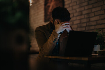 A businessman sits at a desk in a dark office, holding his head in frustration. The dim lighting highlights the stress and pressure of his work environment.