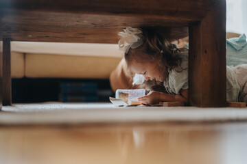 A young girl engrossed in playing and reading underneath a wooden table, exhibiting curiosity and...